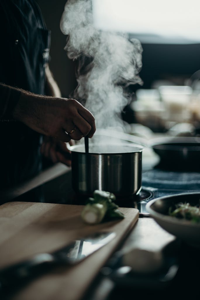 Une personne remuant une casserole fumante sur une cuisinière électrique avec des ustensiles de cuisine étalés.