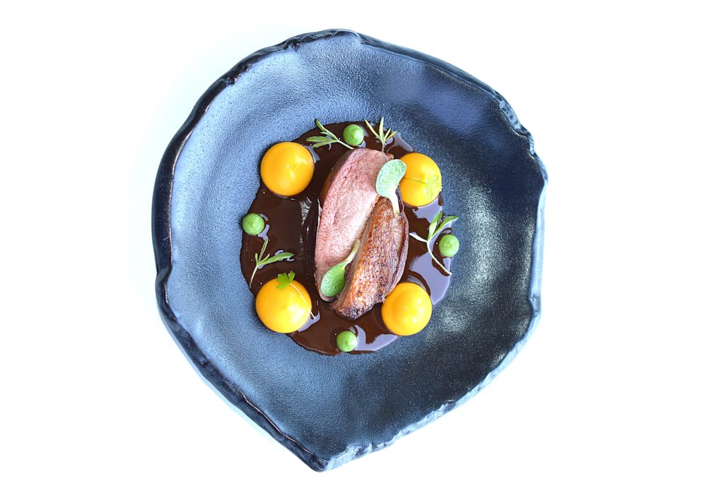 Gourmet dish featuring sliced pigeon breast served on a blue black plate with sauce, yellow purée dots, and microgreens