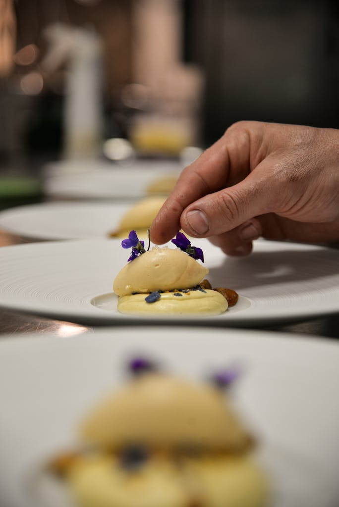 Gros plan sur une main dressant méticuleusement une assiette de dessert gastronomique ornée de fleurs comestibles pour une présentation artistique.