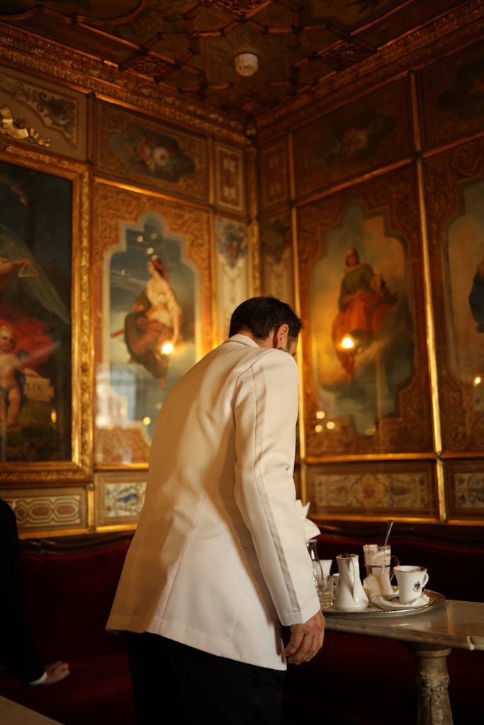 Un serveur en costume blanc à l'intérieur d'un palais opulent et rempli d'œuvres d'art, vu de dos.